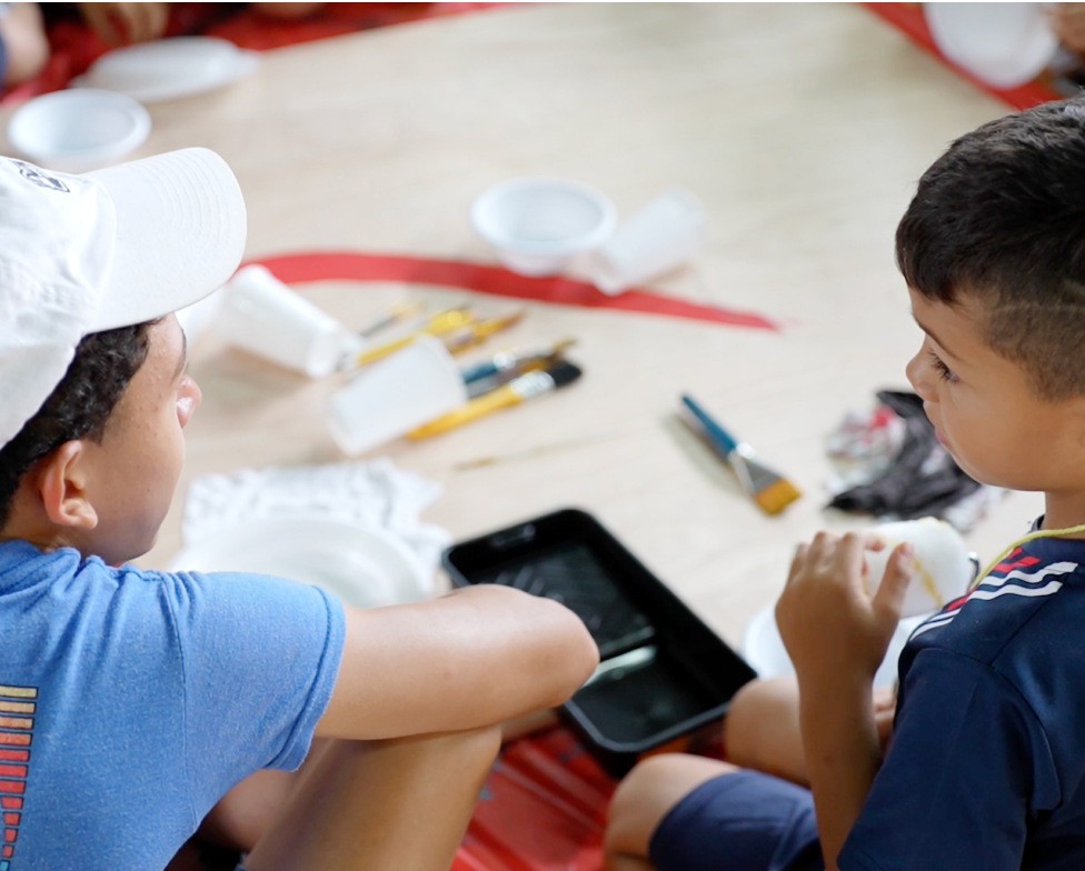 Dos niños, uno con una gorra blanca y el otro con una camiseta azul con rayas rojas, están sentados juntos en el suelo, concentrados en materiales de arte, como pinceles, pinturas y bandejas negras, durante una actividad de pintura.