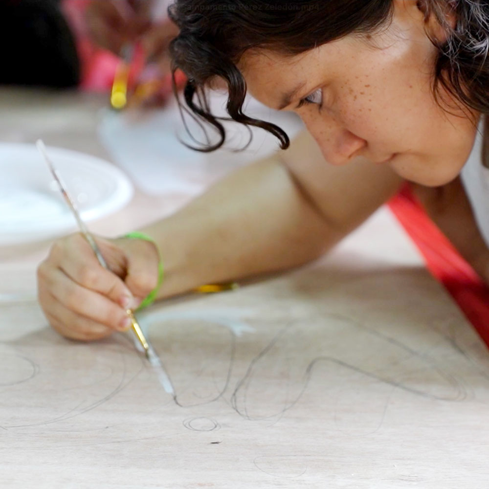 Chica concentrada pintando un gran dibujo a lápiz sobre una superficie de madera clara, utilizando un pincel fino y pintura blanca.