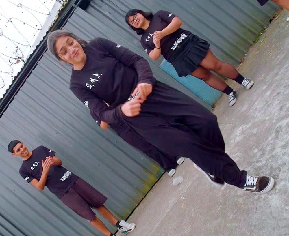 Un grupo de tres jóvenes vestidos de negro ensaya una coreografía de baile en un espacio exterior, con la persona en primer plano realizando un movimiento.